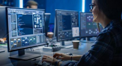 An individual in a plaid shirt is working on a computer at a desk with multiple monitors displaying various graphs and data, in a dark room