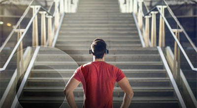 Person wearing headphones standing at the base of a wide staircase, looking upward as sunlight illuminates the steps