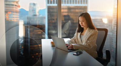 A woman sits at a desk in a modern office with large windows, working on a laptop as sunlight streams in and city buildings are visible outside.