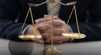 Close-up of a balanced golden justice scale with two pans in focus and clasped hands of an individual in the background