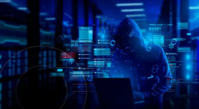 An individual with their face hidden with a hood types on a laptop in a dimly lit server room