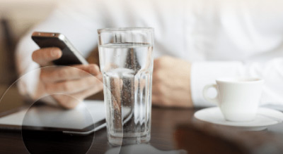 A person’s hand holding a phone with a glass of water and a coffee cup on a wooden table