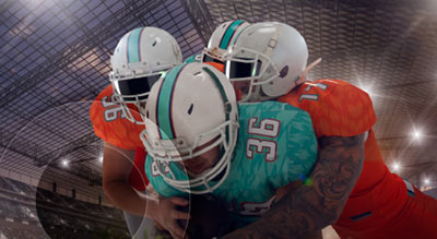 Three football players in teal and orange uniforms collide during a play inside a stadium, with one player being tackled by two defenders