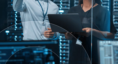 Two people examine a server room while one holds a laptop