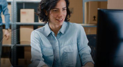 A person in a blue shirt sitting in an office at a desk looking at a computer monitor with boxes in the background