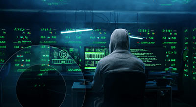 A person in a hoodie sits in front of multiple computer screens displaying green digital data