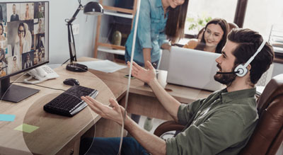 A person is participating in a virtual meeting with multiple people on the screen, while another person is working on a laptop nearby