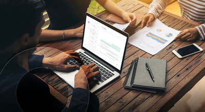A person collaborating with other individuals while working on a laptop with documents