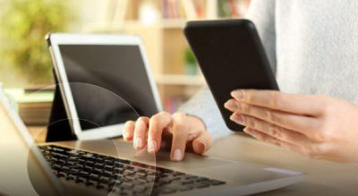 A person is multitasking, using a smartphone in one hand and typing on a laptop with the other in a well-lit room with bookshelves filled with books