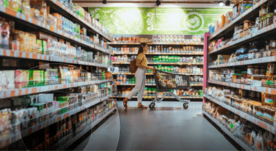  The image captures the inside of a grocery store with fully stocked shelves on both sides of an aisle. A shopper is visible in the middle distance, pushing a shopping cart and browsing products.