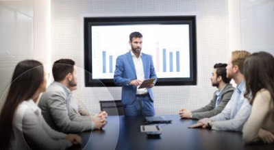 A group of people in a meeting, sitting around a long table with a bar graph presentation on a screen in the background