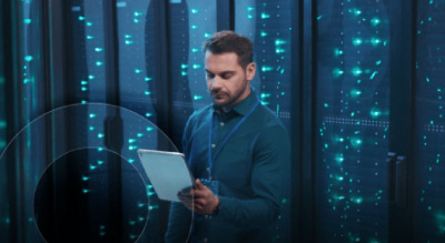 A person in a blue shirt holding a white tablet in front of a black server rack illuminated by blue lights