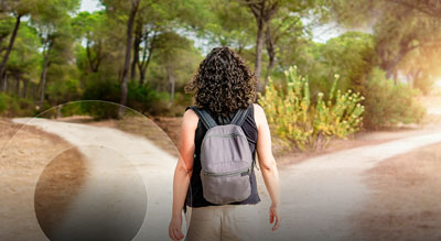 A person with curly hair wearing a backpack walking on a dirt path through a sunlit forest