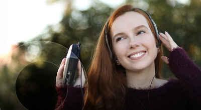 A smiling woman with headphones listens to a cassette player outdoors