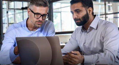 Two individuals in an office setting, one in a blue shirt holding a laptop and the other in a white shirt looking at the laptop