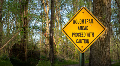 A street sign infront of a wooden area warning of 'Rough Trail Ahead Proceed with Caution'