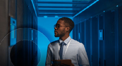 A professional man in a blue shirt and tie stands confidently in a data center, overseeing the technological operations.