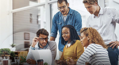 Four people sit at a table looking at a laptop while a fifth person stands behind them; they appear to be working together in a modern office setting.