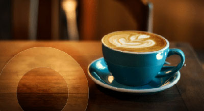 A steaming cup of cappuccino with latte art in a blue cup and saucer, placed on a wooden table next to a round wooden coaster. The coffee appears hot and freshly made, with steam rising from the surfa
