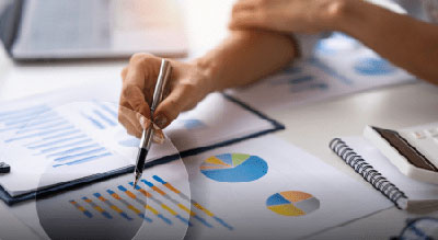 A person is analyzing and pointing at a printed bar graph among various charts and graphs on a desk. Several documents containing different types of data visualizations, including pie charts and bar g