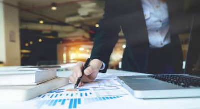 A person’s hand holding a pen over a chart on a desk with a laptop