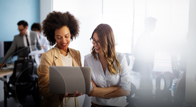 Two individuals are holding and looking at a laptop in a busy office environment