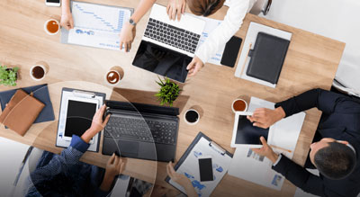 A group of four people working on different devices and documents around a wooden table with coffee and plants.