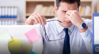 A man sitting at a cluttered desk with a computer monitor, taking off his glasses and pinching the bridge of his nose