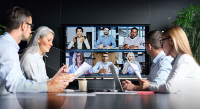 Team members meet around a table with a tv displaying six individuals in a remote call