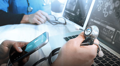 A close-up of a person's hand holding a stethoscope and checking a cellphone in front of a laptop