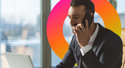 A person in a suit sitting at a desk with a laptop in front of a window while talking on a cellphone