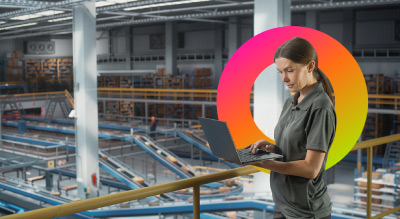 A warehouse or manufacturing facility interior with rows of equipment below, while a worker stands on an elevated walkway using a laptop