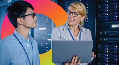 Two individuals stand in front of a server rack, one holding a laptop