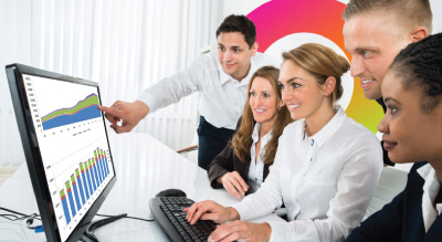 A group of individuals in a meeting around a desk with a computer monitor displaying graphs and charts