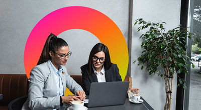 Two individuals collaborate at a table with a laptop, under a vibrant circular wall art in a bright indoor setting