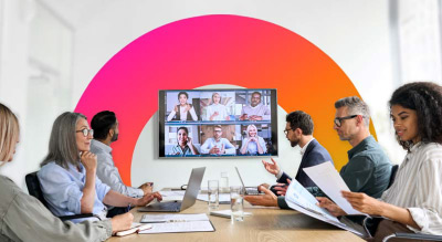 A group of people gathered around a long table with laptops and papers, engaged in a video call displayed on a large screen with six participants