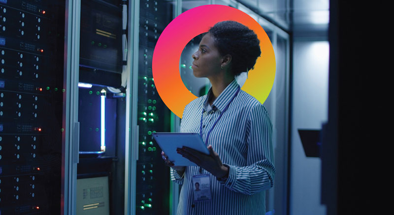A person holding a tablet stands in a server room, inspecting equipment
