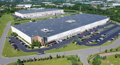 An aerial view of a large industrial building with a parking lot, surrounded by greenery