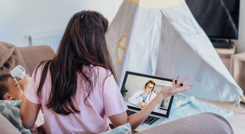 A woman sits on a couch with a laptop on video call with a doctor, while she takes her child's temperature