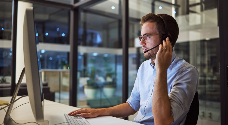 A man in a blue shirt, wearing glasses and a headset, works intently at a computer in a modern office, surrounded by glass walls and dim lighting.
