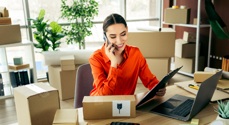 A woman in a bright orange shirt smiles while on the phone at a desk, surrounded by cardboard boxes and a laptop.