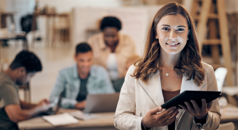 A smiling woman in focus holds a tablet in a modern office, with three colleagues blurred in the background working collaboratively at a table.