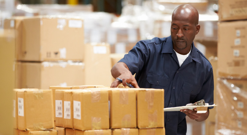 A warehouse worker in a navy shirt inspects stacked cardboard boxes, holding a clipboard.