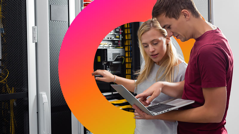 Two people examine servers while using a laptop