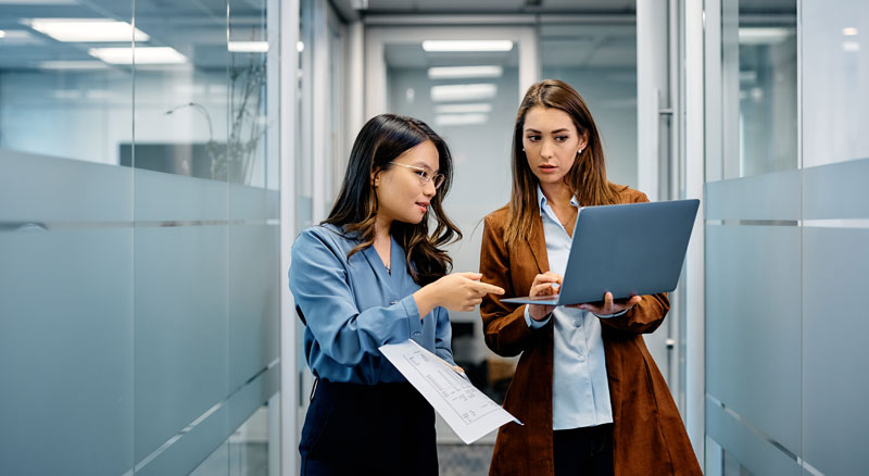 Two individuals discussing a document and working on a laptop in a modern office corridor