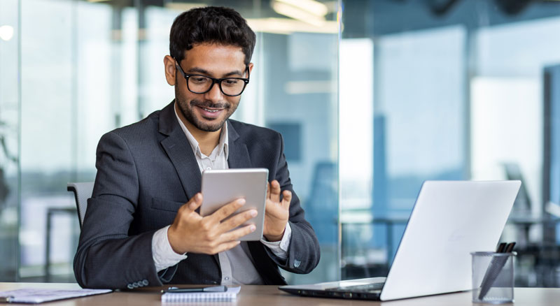 A professional in a suit holds a tablet while sitting at a desk with an open laptop in a modern office setting