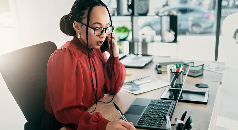 A woman talking on a cellphone writes note infront of her laptop