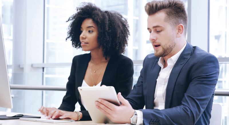Two business professionals seated at a desk, focused on a tablet in front of them