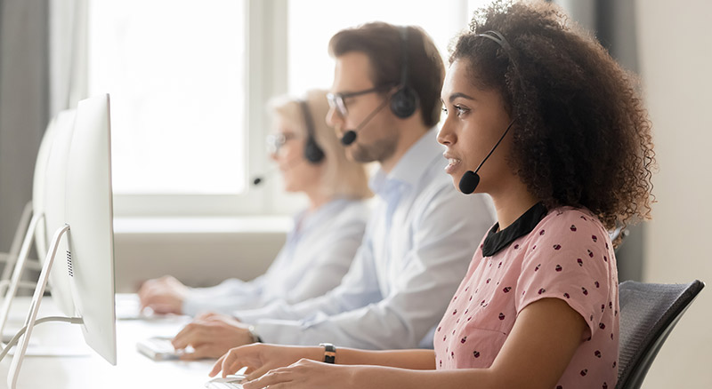 Three individuals with obscured faces are wearing headsets and working at computers in a bright office setting