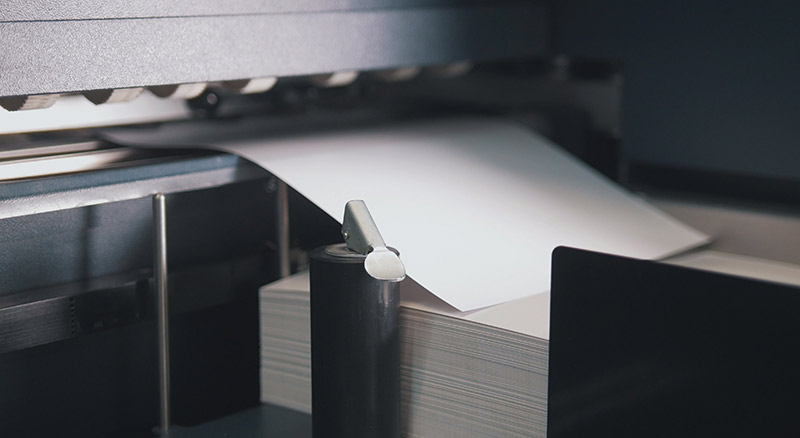 A close-up view of a white sheet of paper emerging from an industrial printing press, with a stack of printed papers below